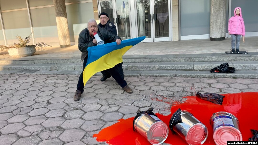 Жандармерист избутва фотографа Рафаело Казаков, който държи украинското знаме пред входа на руското консулство в София. На преден план - червена боя, която символизира кръвта на украински деца. В розово - манекен с фигура на момиче.