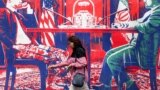 A woman walks past a mural depicting an Iranian official in civilian clothing sitting across the table from an American official wearing military fatigues painted on the outer walls of the former US Embassy in Tehran.