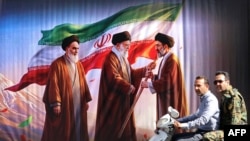 Burra kalojnë me skuter pranë një billbordi ku shfaqen themeluesi i Revolucionit Islamik dhe udhëheqësi i parë suprem, ajatollah Ruhollah Khomeini (majtas), pasardhësi i tij Ali Khamenei (në qendër) dhe djali i këtij të fundit, udhëheqësi aktual suprem, Mojtaba Khamenei.