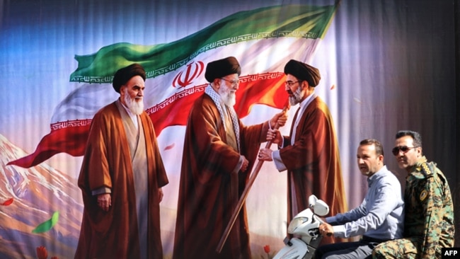 Burra kalojnë me skuter pranë një billbordi ku shfaqen themeluesi i Revolucionit Islamik dhe udhëheqësi i parë suprem, ajatollah Ruhollah Khomeini (majtas), pasardhësi i tij Ali Khamenei (në qendër) dhe djali i këtij të fundit, udhëheqësi aktual suprem, Mojtaba Khamenei.