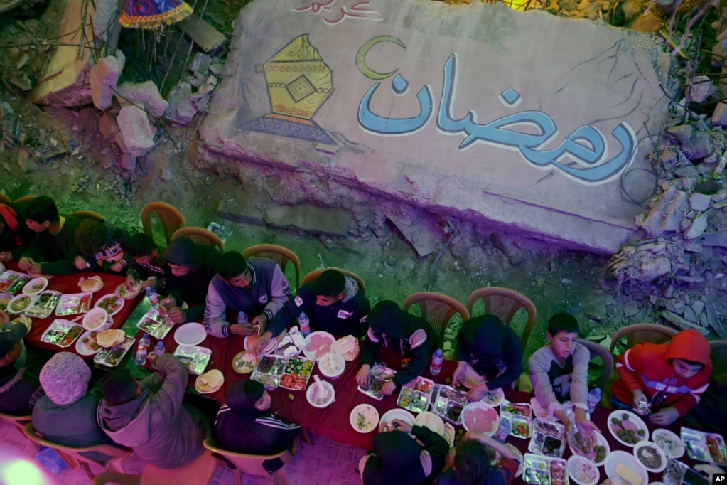 Mbishkrimi "Ramazan", i shkruar arabisht. Palestinezët duke ngrënë syfyrin e parë të shtruar në mënyrë kolektive pranë shtëpive të tyre të shkatërruara në Han Jounis, në jug të Rripit të Gazës, të premten, 28 shkurt 2025.&nbsp;