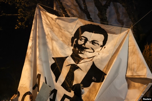 Një person duke ecur afër banerit me fotografinë e kryetarit të Stambollit, Ekrem Imamoglu në një protestë kundër ndalimit të tij.