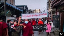 Fiatal aktivisták és buddhista szerzetesek vonulnak a hadsereg ellen tiltakozva Mandalajban. A transzparensen az áll, hogy „Ki merészel az emberek akaratával ellentétes oldalon állni?” – 2022. február 1.