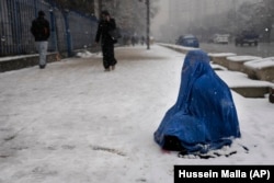 Një grua afgane me burka, e ulur në borë, duke kërkuar lëmoshë. Kabul 2022.