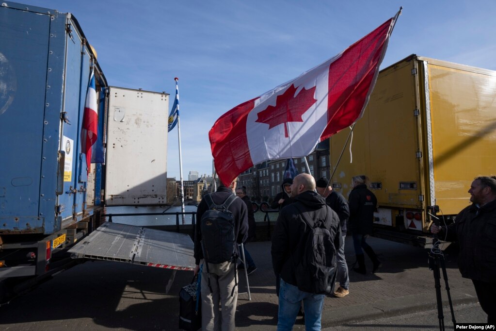 Demonstruesit - njëri prej tyre me një flamur kanadez - duke qëndruar pranë disa kamionëve që bllokuan një hyrje në ndërtesat qeveritare, në Hagë të Holandës. Ata bënin thirrje kundër masave anti-COVID. (AP)
