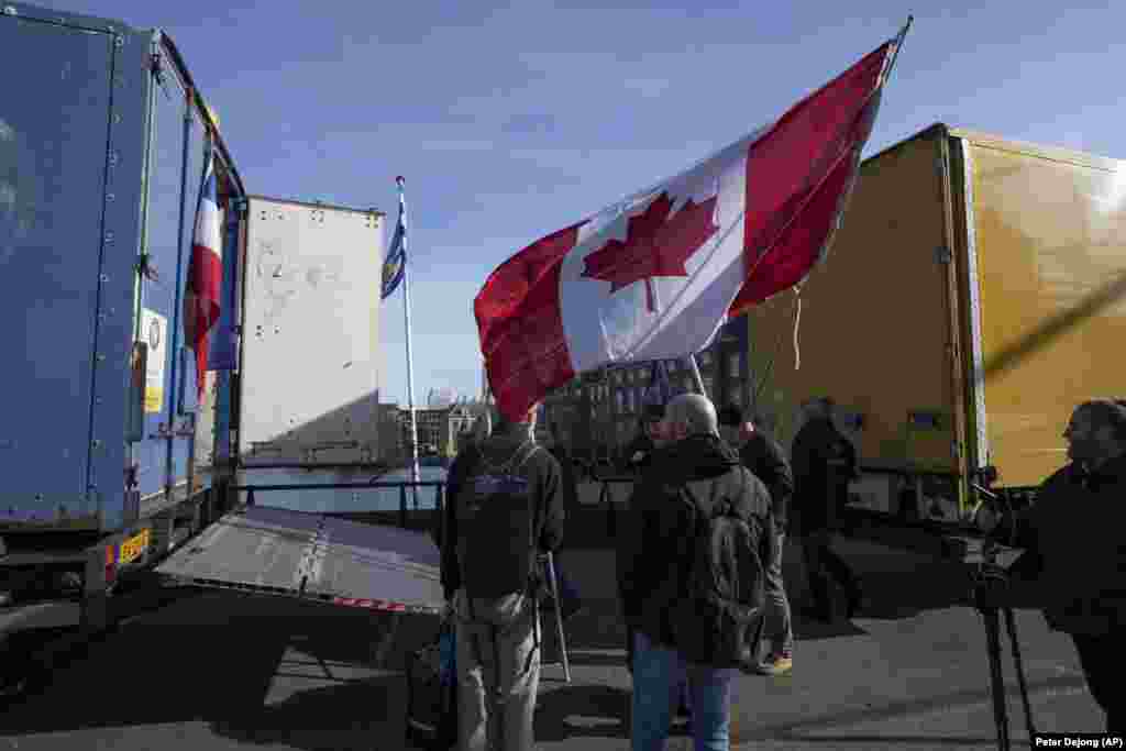 Демонстранти стоять поруч із близько 20 вантажівками, які заблокували один із входів до урядових будівель в Гаазі, на знак протесту проти обмежень пов&#39;язаних із COVID-19. Нідерланди. 12 лютого 2022 року. Один із протестувальників тримає в руках канадський прапор. Людей в Європі на протести частково надихнули якраз протести в Канаді