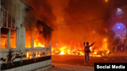 Протестиращ в Иран вдига ръце във въздуха на фона на горящи коли и сгради, 8 януари 2026 г.