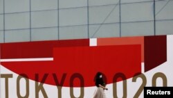 Arcmaszkot viselő nő sétál a nyári olimpia logója előtt 2021. július 29-én Tokióban