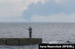 Чоловік ловить рибу з пірсу в Одесі на тлі пожежі в порту Південний