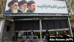 Иранцы сидят на автобусной остановке под билбордом с изображениями покойного верховного лидера Ирана аятоллы Рухоллы Хомейни (слева), его преемника, покойного аятоллы Али Хаменеи (в центре), и нового верховного лидера Моджтабы Хаменеи в Тегеране, 31 марта 2026 года