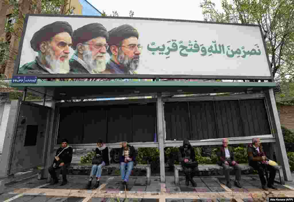 Iranians sit at a bus stop in Tehran on March 31.The station features a billboard depicting (left to right) Iran's ayatollahs Ruhollah Khomeini, who died in 1989, Ali Khamenei, who was killed in US-Israeli strikes on February 28, and Mojtaba Khamenei, the current ruler of the country.