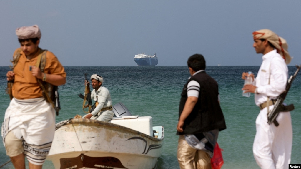 گروهی از مردان مسلح حوثی‌ مسلح در ساحل؛ در نمای دورتر کشتی تجاری گلکسی لیدر دیده می‌شود که توسط آن‌ها توقیف شده است