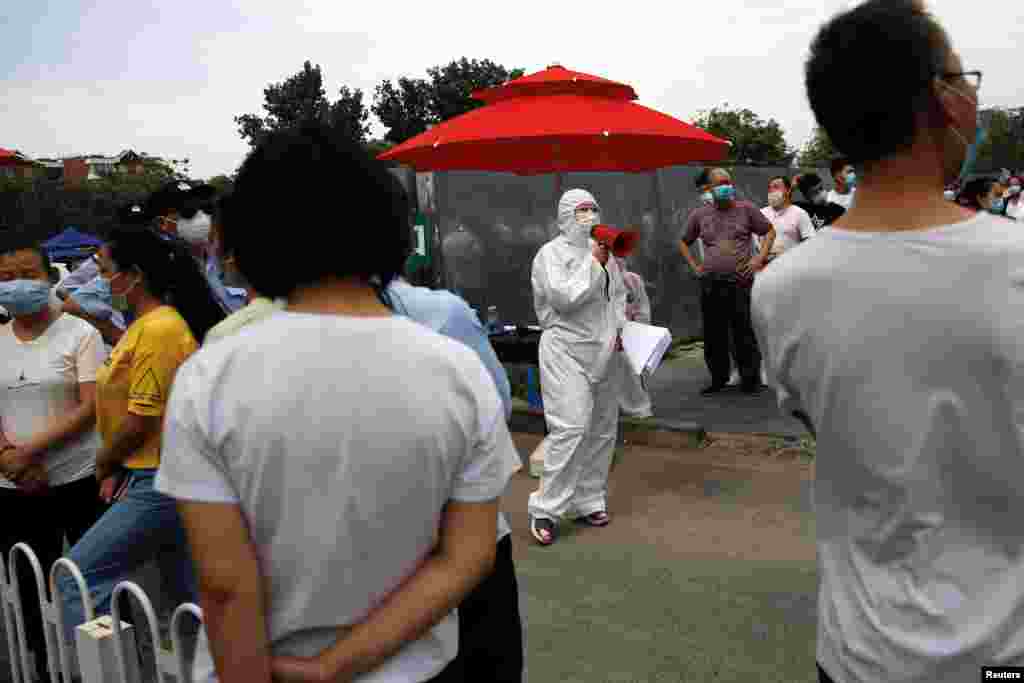 Медицинский работник в защитном костюме с громкоговорителем обращается к людям, которые выстроились в очередь для сдачи анализов на коронавирус