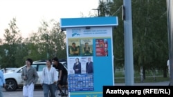 Сайлау алдындағы үгіт-насихат қағаздары ілінген стендтің жанынан өтіп бара жатқан жастар. Астана 2026 жылғы шілде.