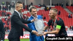 Középen Bánki Erik fideszes képviselő segít Szijjártó külügyminiszternek, hogy átadja a másodosztályú bajnoki serleget Dzsudzsák Balázs labdarúgónak Debrecenben 2021. május 1-jén