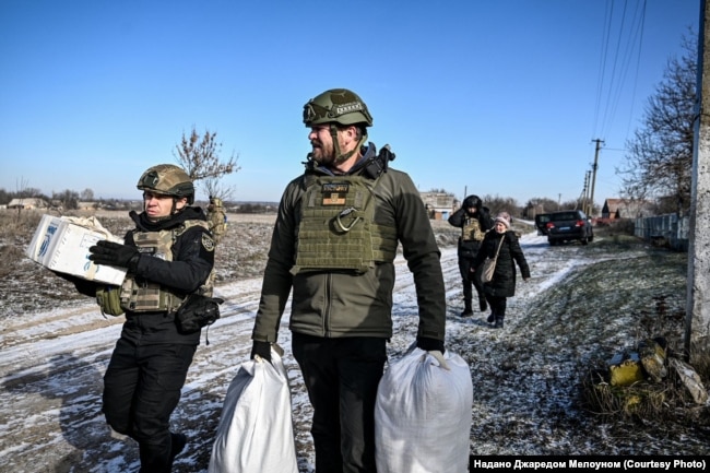 Джаред Мелоун разом поліційським несуть до евакуаційного транспорту речі людей, які виїздять з прифронтового села, Запорізька область, лютий 2026 року
