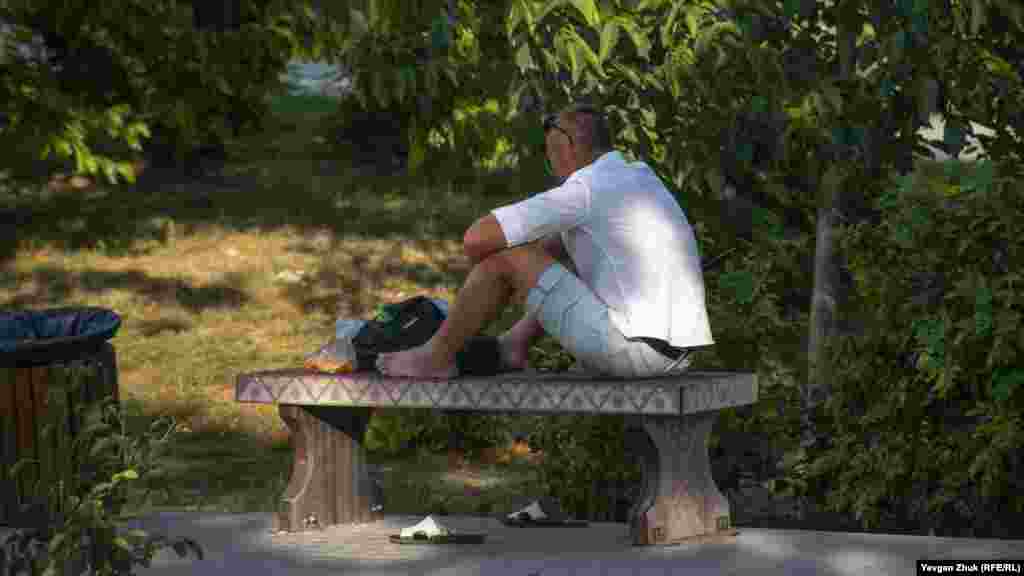 Мужчина отдыхает на лавке в тени деревьев