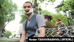 Грузинские печатные издания, помещенные в клетку, и человек, прикованный к ней наручниками, люди с белыми повязками, закрывающими глаза и рты