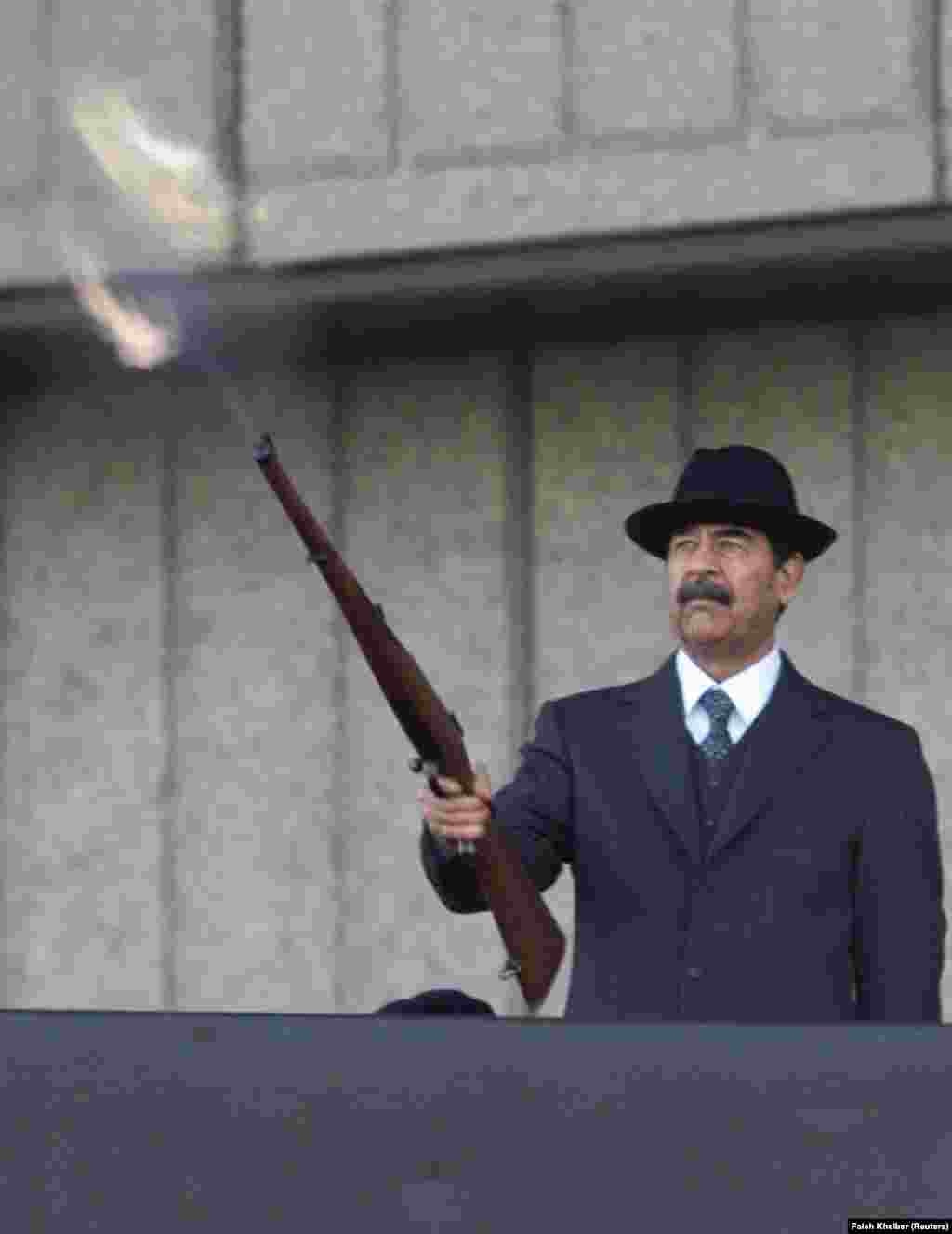 Президент Ирака Саддам Хусейн стреляет из винтовки, которую держит в одной руке. В другой &mdash; сигара. Действие происходит во время военного парада в декабре 2000 года.