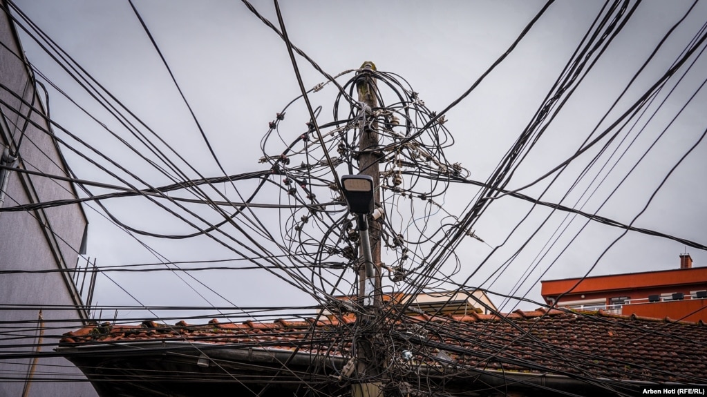 Tela të rrymës, kabllo të shumta telefonike, interneti e të tjera, në një shtyllë betoni që pothuajse mbulojnë kulmin e një shtëpie në lagjen "Vellusha" në kryeqytet. 