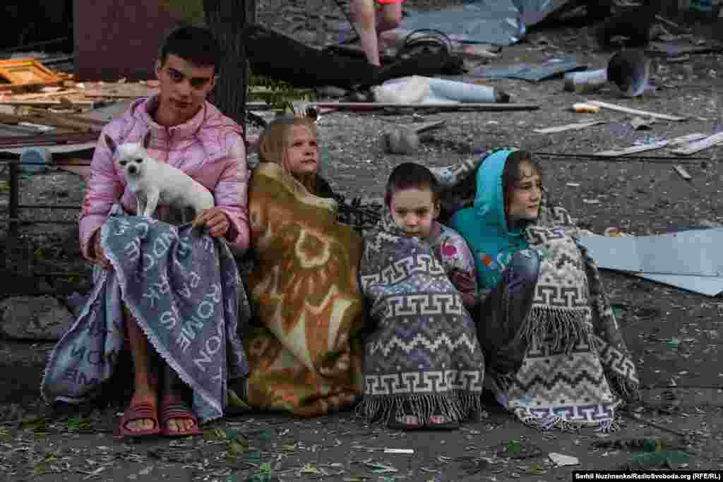 Діти, закутані у ковдри, сидять разом із своїм песиком біля свого будинку, пошкодженого внаслідок російської атаки. Серпень 2025 року