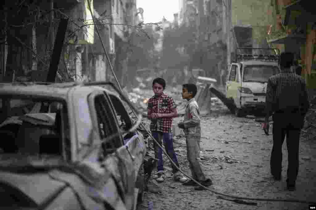 21 жовтня 2015 року. Сирійські діти з передмістя Дамаска стоять біля уламків, що залишилися після авіаударів по територіях, які утримуються опозиційними до режиму Башара Асада збройними формуваннями. 30 вересня 2015 року Росія почала свою повітряну кампанію у Сирії і таким чином долучилася до громадянської війни на стороні Асада. США і НАТО заявляють, що російські літаки бомблять позиції поміркованої опозиції у Сирії, які воюють проти угруповання &laquo;Ісламська держава&raquo;. Російська сторона заявляє, що всі групи, які воюють проти урядових сил у Сирії, є терористичними