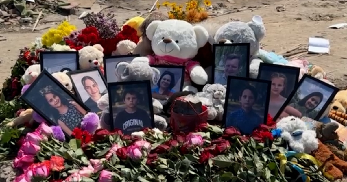 A memorial arrangement featuring framed photos, teddy bears, and flowers on sand.