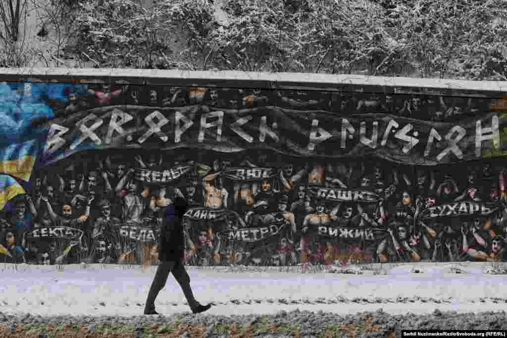 На фото: чоловік проходить повз мурал на підпірній стіні в Протасовому Яру, Київ, лютий 2026 року.Цей мурал створений на основі фотографії, зробленої під час футбольного матчу між «Динамо» Загреб та «Хайдук» Спліт у лютому 2023 року у Загребі (Хорватія). Коли вболівальники «Бед Блю Бойз» з «Динамо» Загреб вшанували пам'ять українських військових – футбольних уболівальників, які загинули в боях з російськими військовими, – піднявши вгору руки з банерами, на яких були зображені імена загиблих та гасла «Слава Україні!»