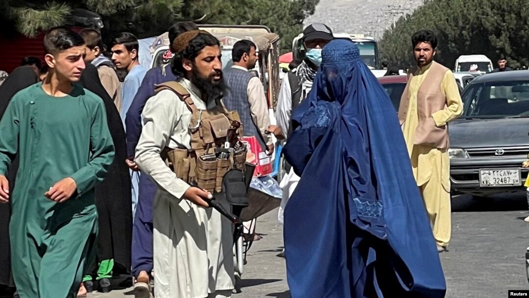 Forțele talibane blochează drumurile din jurul aeroportului, în timp ce o femeie îmbrăcată în Burqa trece pe lângă, Kabul, Afganistan. Fotografie realizată în 27 august 2021.